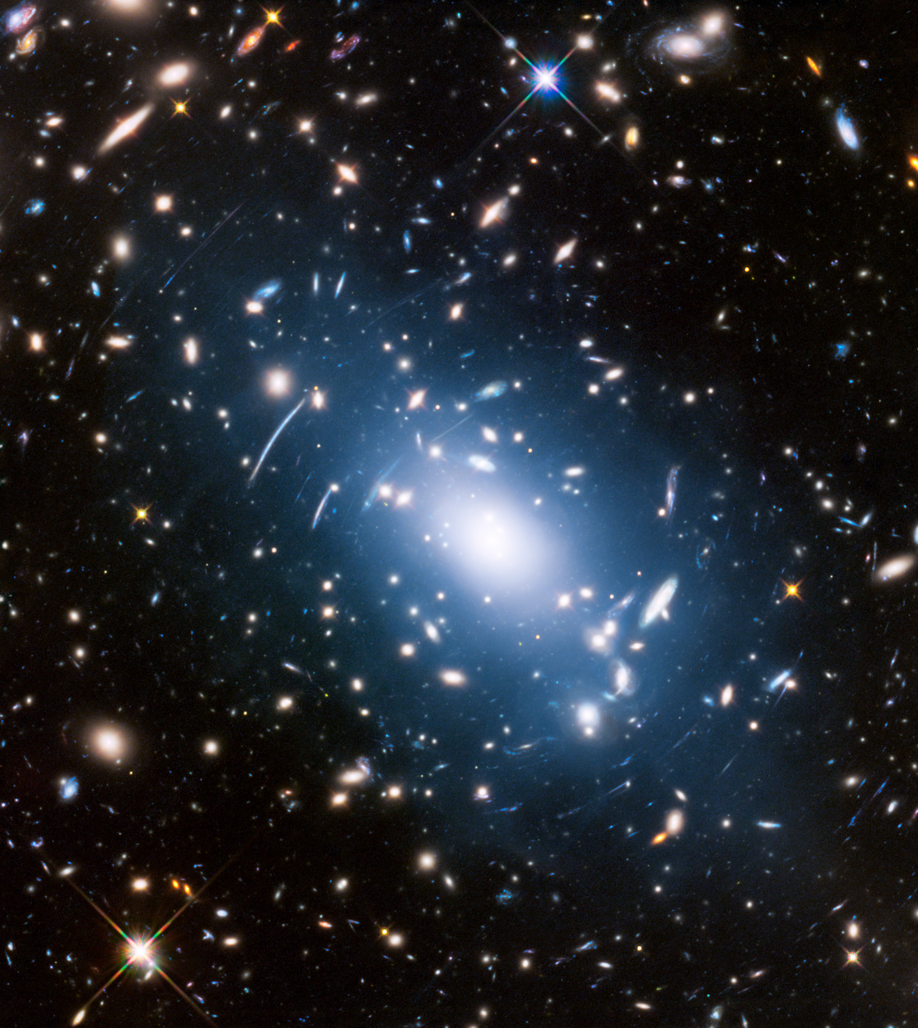 Massive galaxy cluster Abell S1063 is shown at the center of this Hubble image, surrounded by more distant galaxies that are magnified and warped by the cluster’s immense gravity.  A faint haze of intracluster light is visible between the galaxies, produced by free-floating stars.