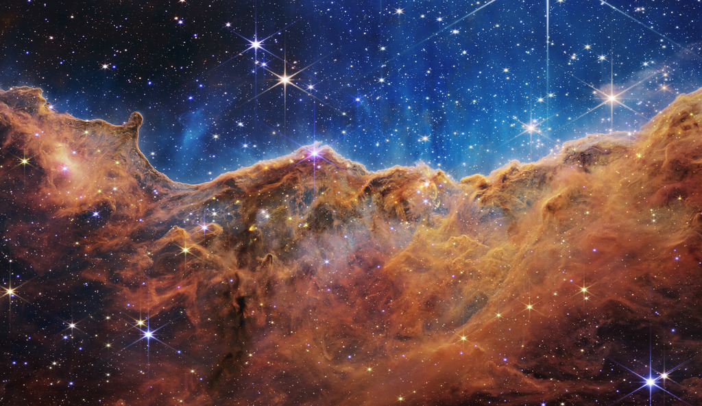 The seemingly three-dimensional “Cosmic Cliffs” showcases Webb’s capabilities to peer through obscuring dust and shed new light on how stars form. Webb reveals emerging stellar nurseries and individual stars that are completely hidden in visible-light pictures. This landscape of “mountains” and “valleys” is actually the edge of a nearby stellar nursery called NGC 3324 at the northwest corner of the Carina Nebula.So-called mountains — some towering about 7 light-years high — are speckled with glittering, young stars imaged in infrared light. A cavernous area has been carved from the nebula by the intense ultraviolet radiation and stellar winds from extremely massive, hot, young stars located above the area shown in this image. The blistering, ultraviolet radiation from these stars is sculpting the nebula’s wall by slowly eroding it away. Dramatic pillars rise above the glowing wall of gas, resisting this radiation. The “steam” that appears to rise from the celestial “mountains” is actually hot, ionized gas and hot dust streaming away from the nebula due to the relentless radiation.Objects in the earliest, rapid phases of star formation are difficult to capture, but Webb’s extreme sensitivity, spatial resolution and imaging capability can chronicle these elusive events.